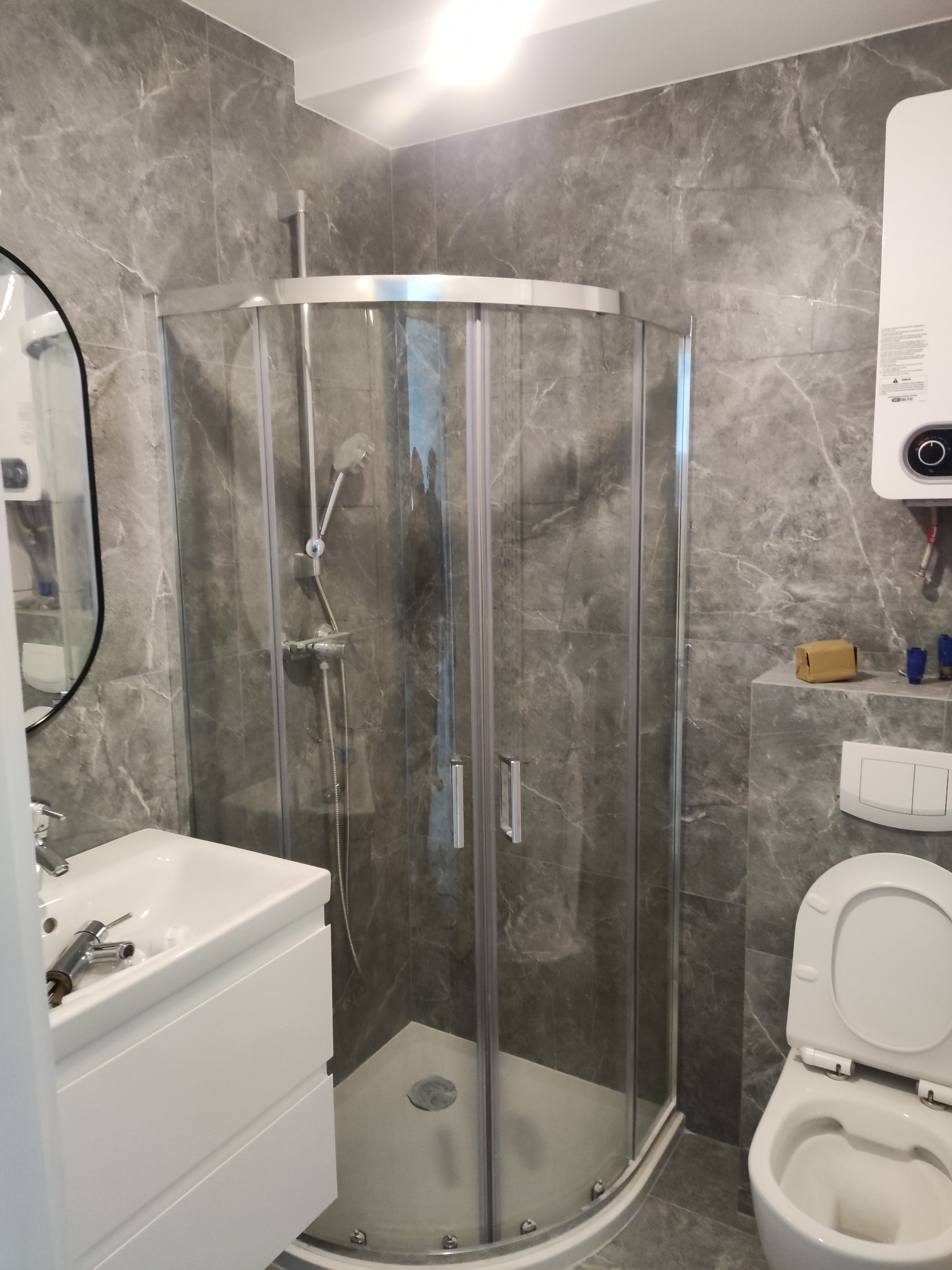 Nowoczesna łazienka z szarymi marmurkowymi płytkami, kabiną prysznicową z zaokrąglonymi drzwiami, białą umywalką z szafką i otwartą toaletą. Widoczne narzędzia na umywalce sugerują trwający montaż.