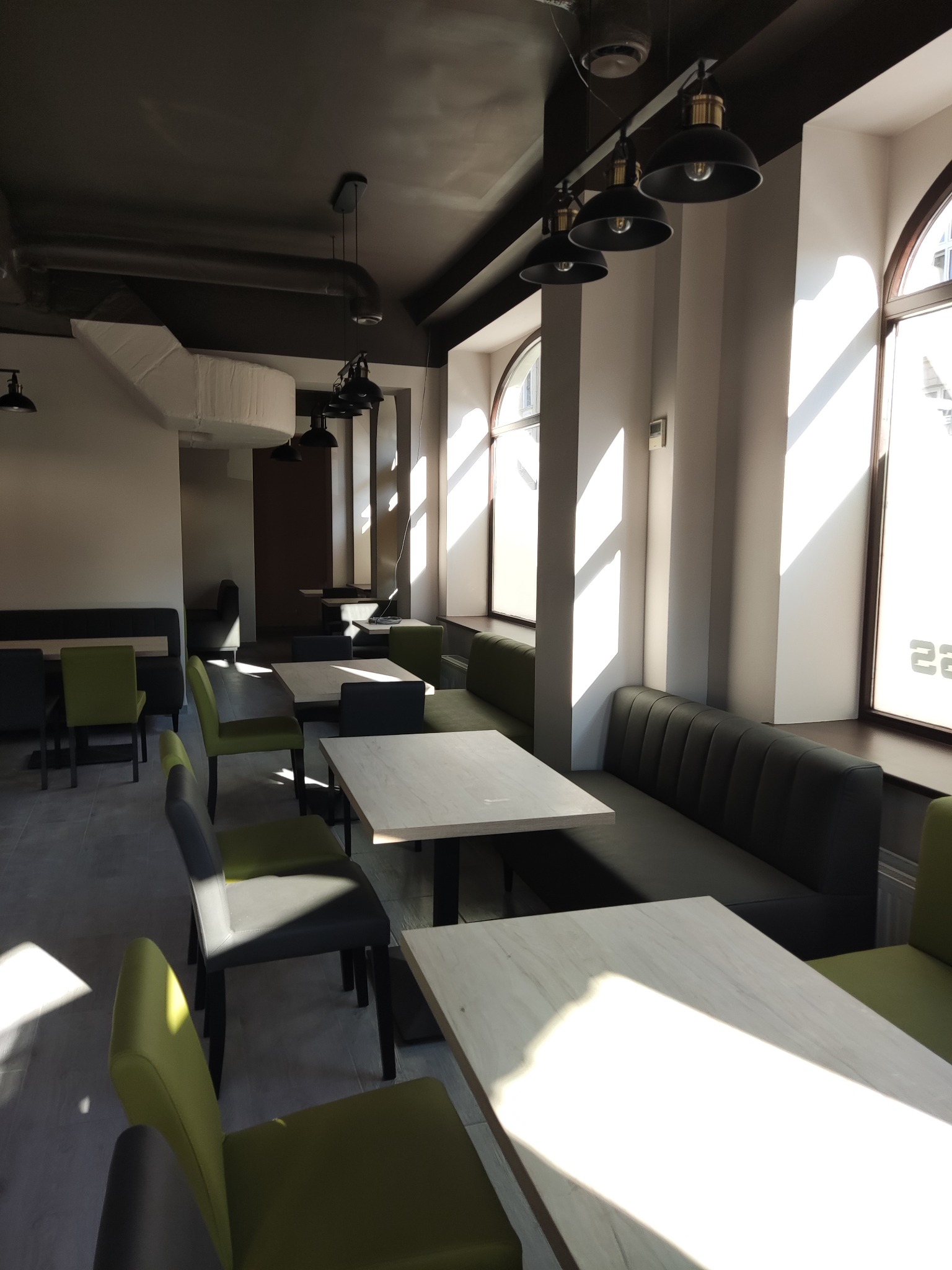 Jasne wnętrze kawiarni lub restauracji z rzędami okien i wpadającym przez nie światłem słonecznym, ciemne sufity z lampami w stylu industrialnym, stoły z jasnymi blatami i krzesłami w odcieniach...