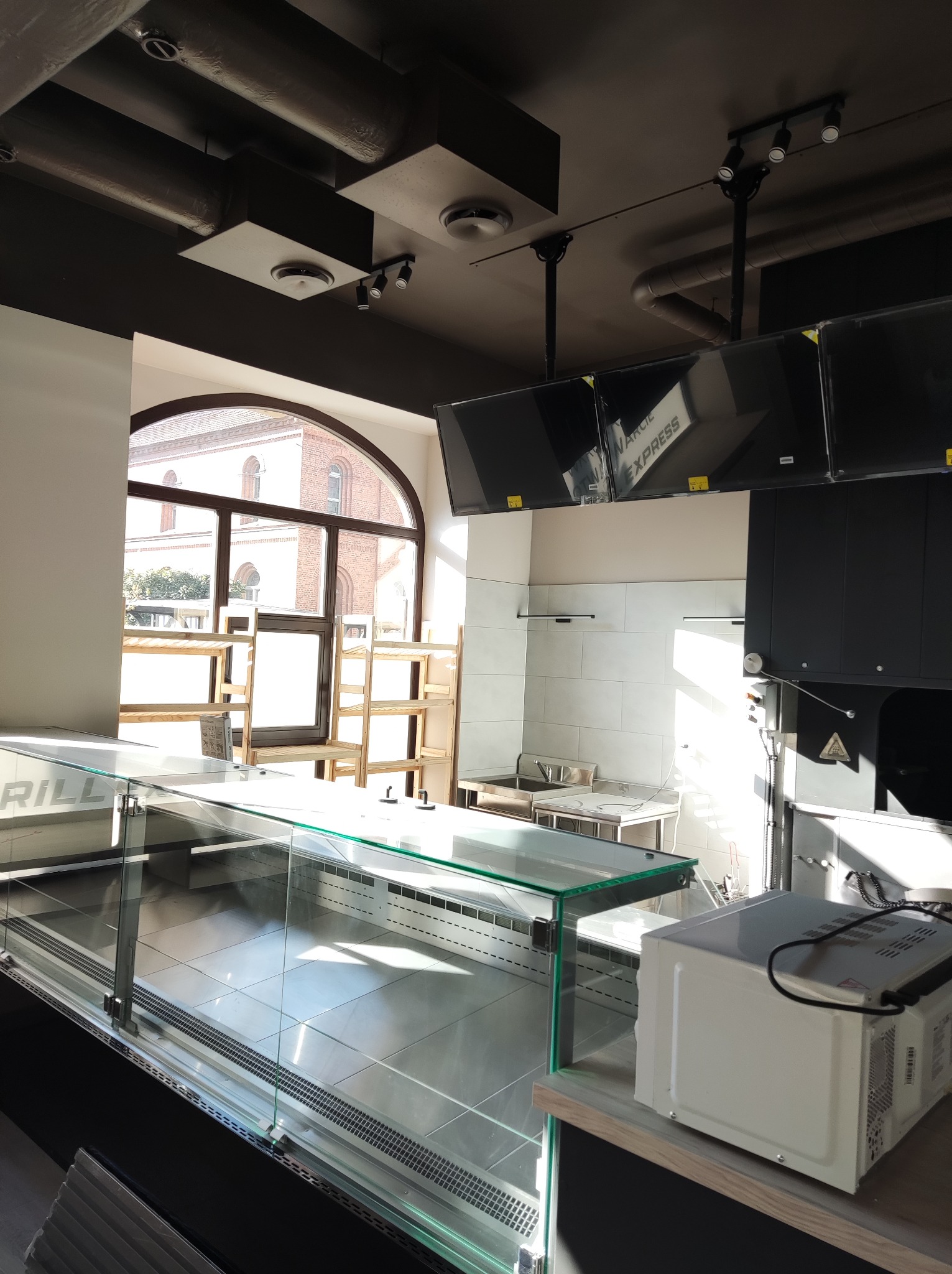 Wnętrze lokalu gastronomicznego w trakcie wykańczania: widoczna przeszklona lada chłodnicza, białe płytki na ścianie, surowe drewniane regały pod oknem, oraz instalacje pod sufitem w kolorze...