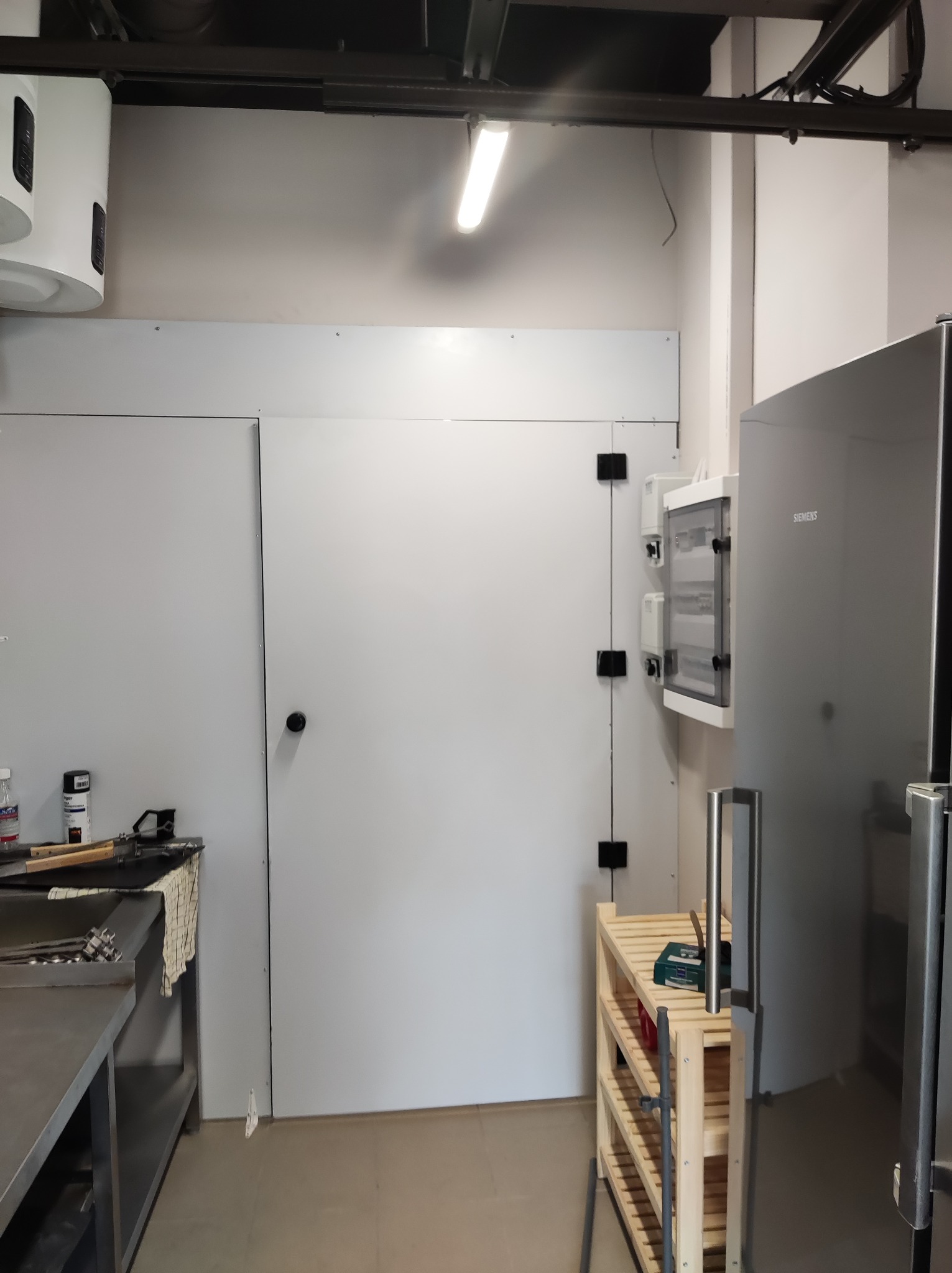 Wnętrze pomieszczenia z zabudową z płyt kartonowo-gipsowych, widoczne szafki elektryczne, bojler, metalowy blat i regał drewniany.