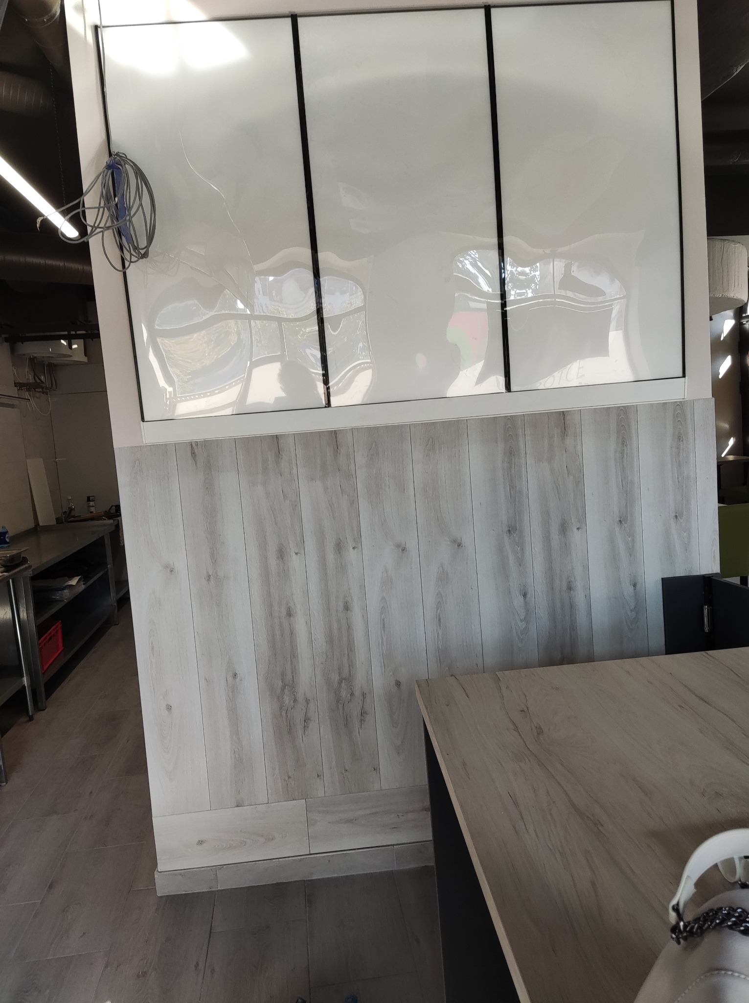 Ściana wykończona panelami drewnopodobnymi z białymi, błyszczącymi płytami na górze, rozdzielonymi czarnymi listwami. Widoczne kable i fragment stołu.