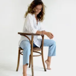 Kobieta z kręconymi włosami, ubrana w białą koszulę i jasne dżinsy, siedzi bokiem na drewnianym krześle na białym tle, spoglądając w dół z uśmiechem.
