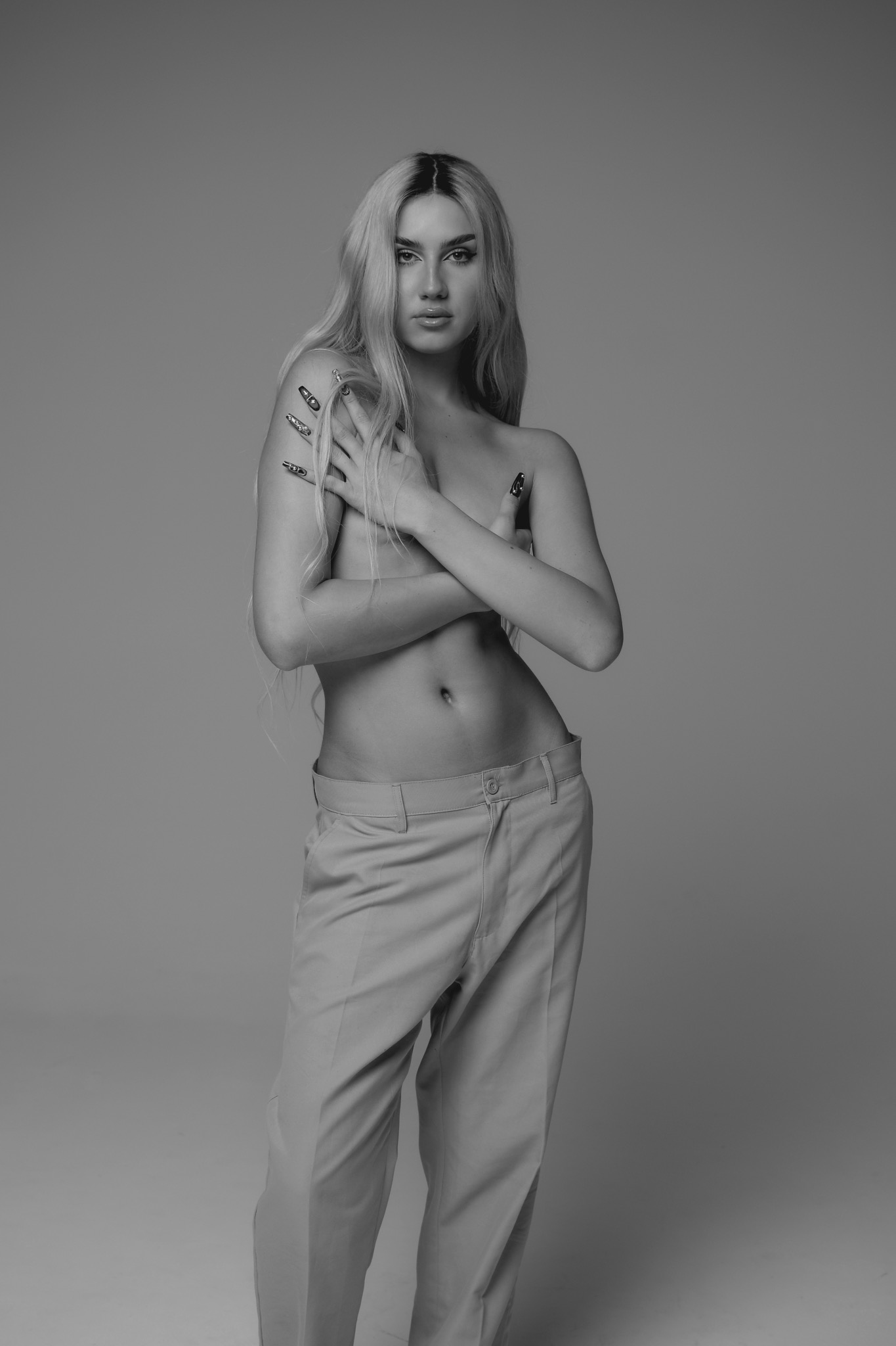 Czarno-białe portretowe ujęcie kobiety z długimi blond włosami, pozującej w spodniach i z ramionami skrzyżowanymi na piersiach, z widocznymi tatuażami na ramieniu i oryginalnym manicure.
