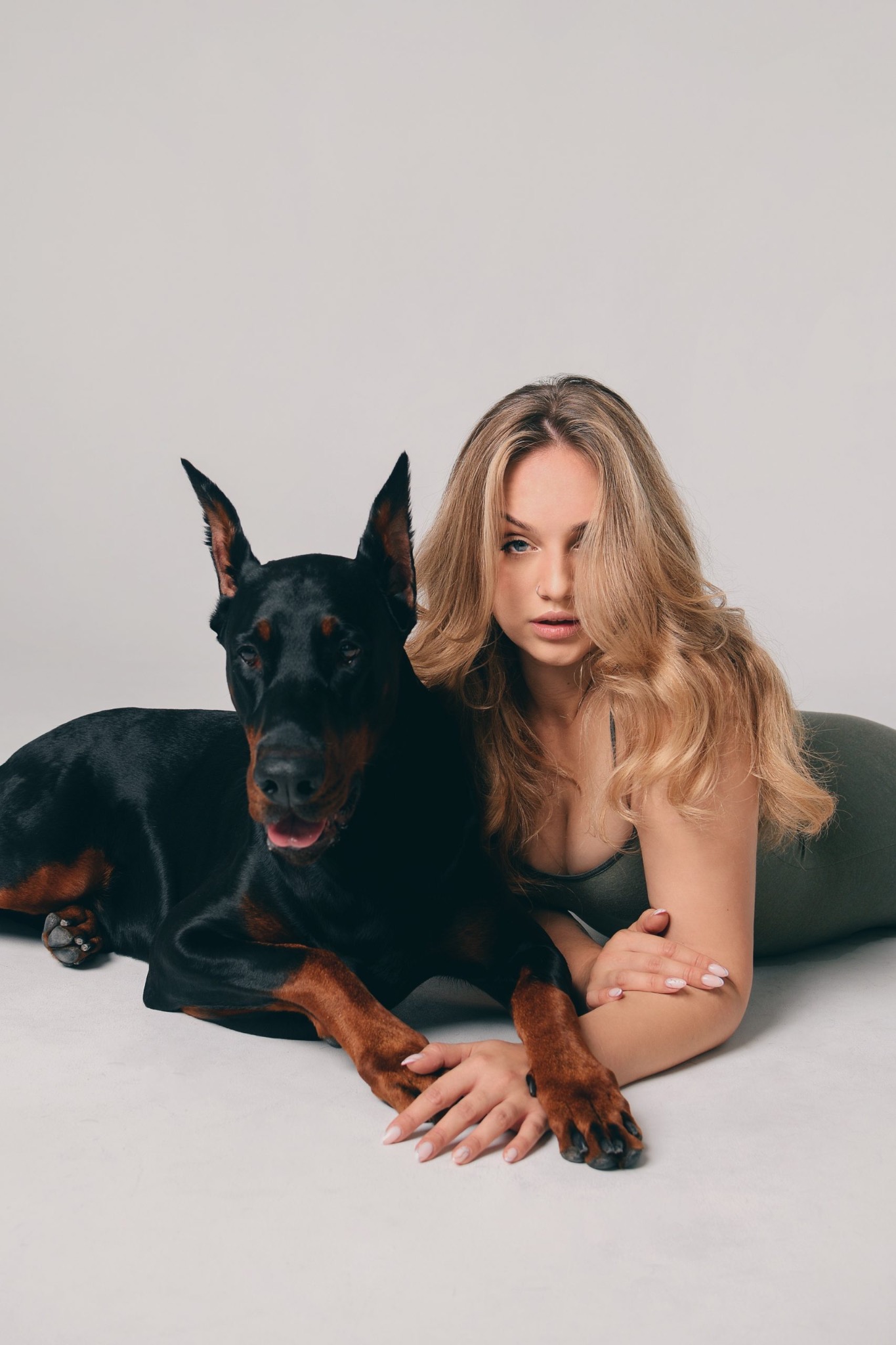 Portret kobiety z blond włosami leżącej obok czarnego dobermana na jasnym tle, z delikatnym makijażem i nose ringiem.