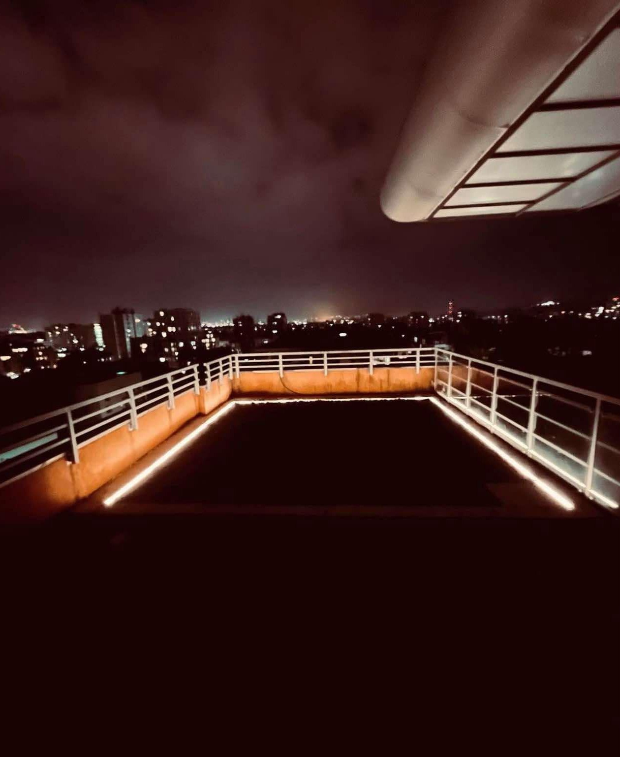 Nocny widok z tarasu na dachu budynku z metalową balustradą i subtelnym oświetleniem LED wzdłuż krawędzi, w tle panorama miasta rozświetlona licznymi światłami.