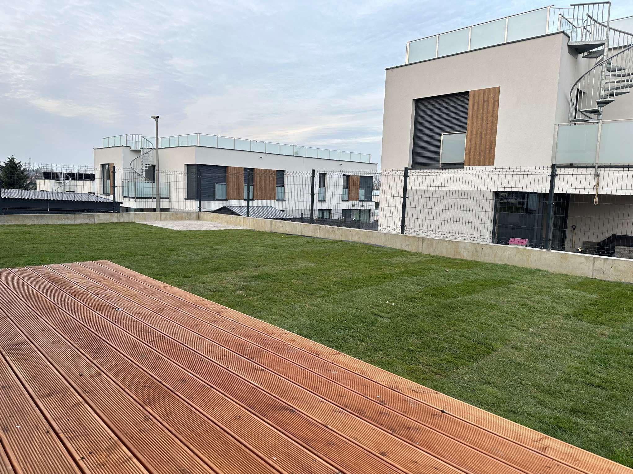 Widok na świeżo założony trawnik z rolki na dachu budynku mieszkalnego, z drewnianym tarasem na pierwszym planie i nowoczesną architekturą w tle.