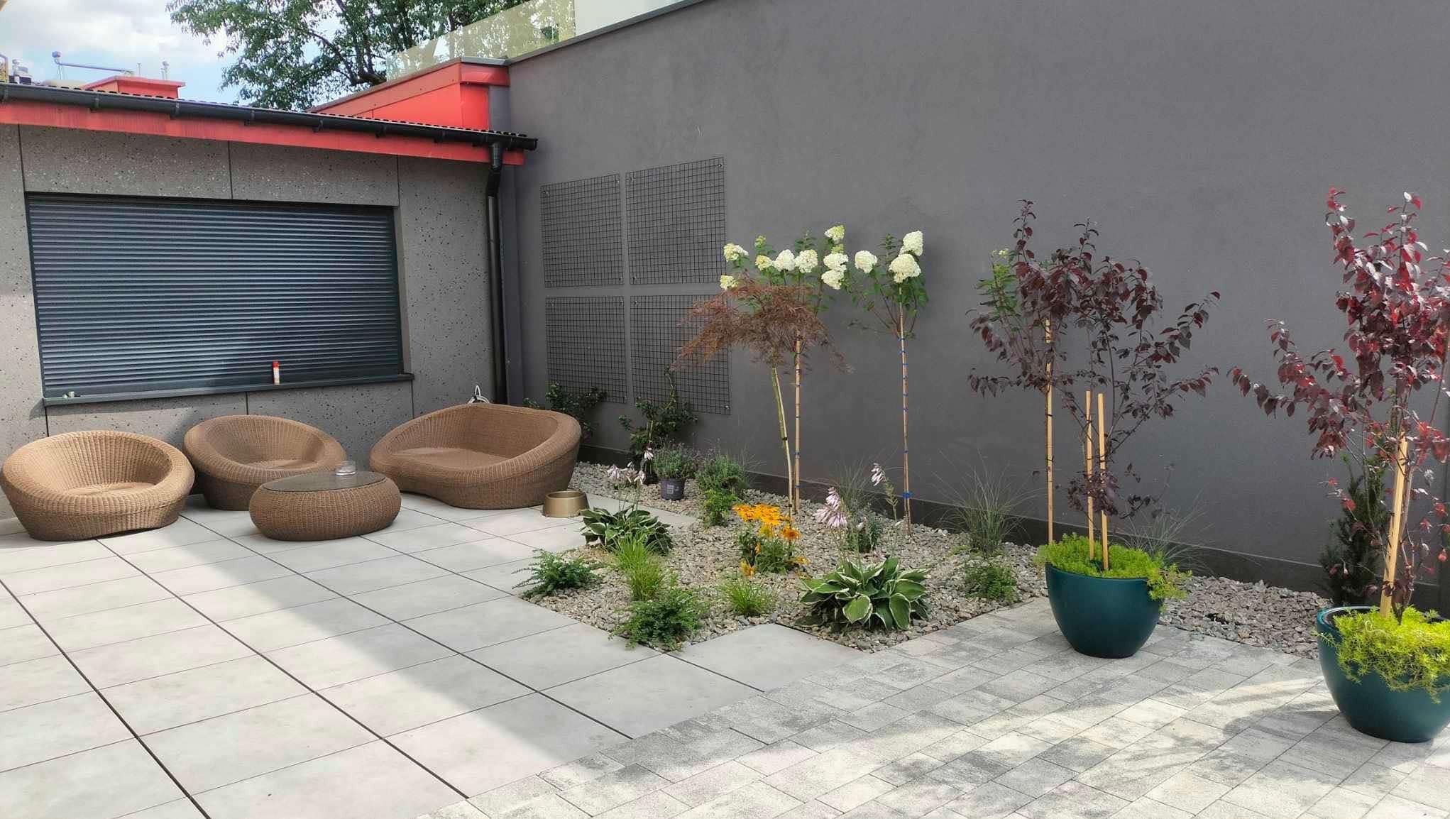 Nowoczesny taras z szarymi płytami, rattanowymi meblami wypoczynkowymi i rabatą z kamyków, drzewek i kwiatów. Dwa drzewka w donicach. Ściana w tle z kratkami.