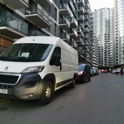 Biały samochód dostawczy marki Peugeot z kartką informacyjną na przedniej szybie, zaparkowany na tle nowoczesnych budynków mieszkalnych z balkonami.