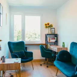 Jasny gabinet psychoterapeuty z dwoma turkusowymi fotelami, biurkiem, regałem z książkami i słonecznikami w wazonie, widok z okna.
