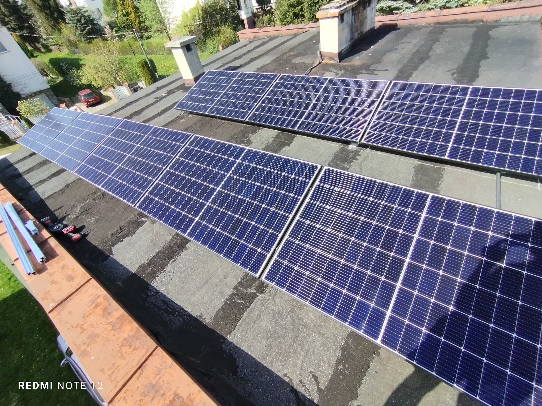 Instalacja paneli słonecznych na płaskim dachu pokrytym papą, widoczne narzędzia i profile montażowe, budynki w tle.