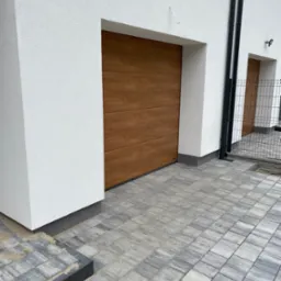 Nowoczesna brama garażowa segmentowa w kolorze drewna, z poziomymi panelami, wkomponowana w białą elewację budynku z kostki brukowej na podjeździe.