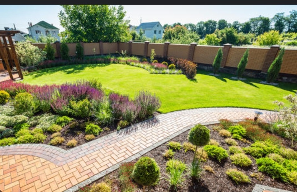 Ujęcie z góry na zadbany ogród z równo przystrzyżonym trawnikiem, rabatami kwiatowymi z fioletowymi i zielonymi roślinami, ścieżką z cegły oraz wysokim, ceglanym murem w tle.