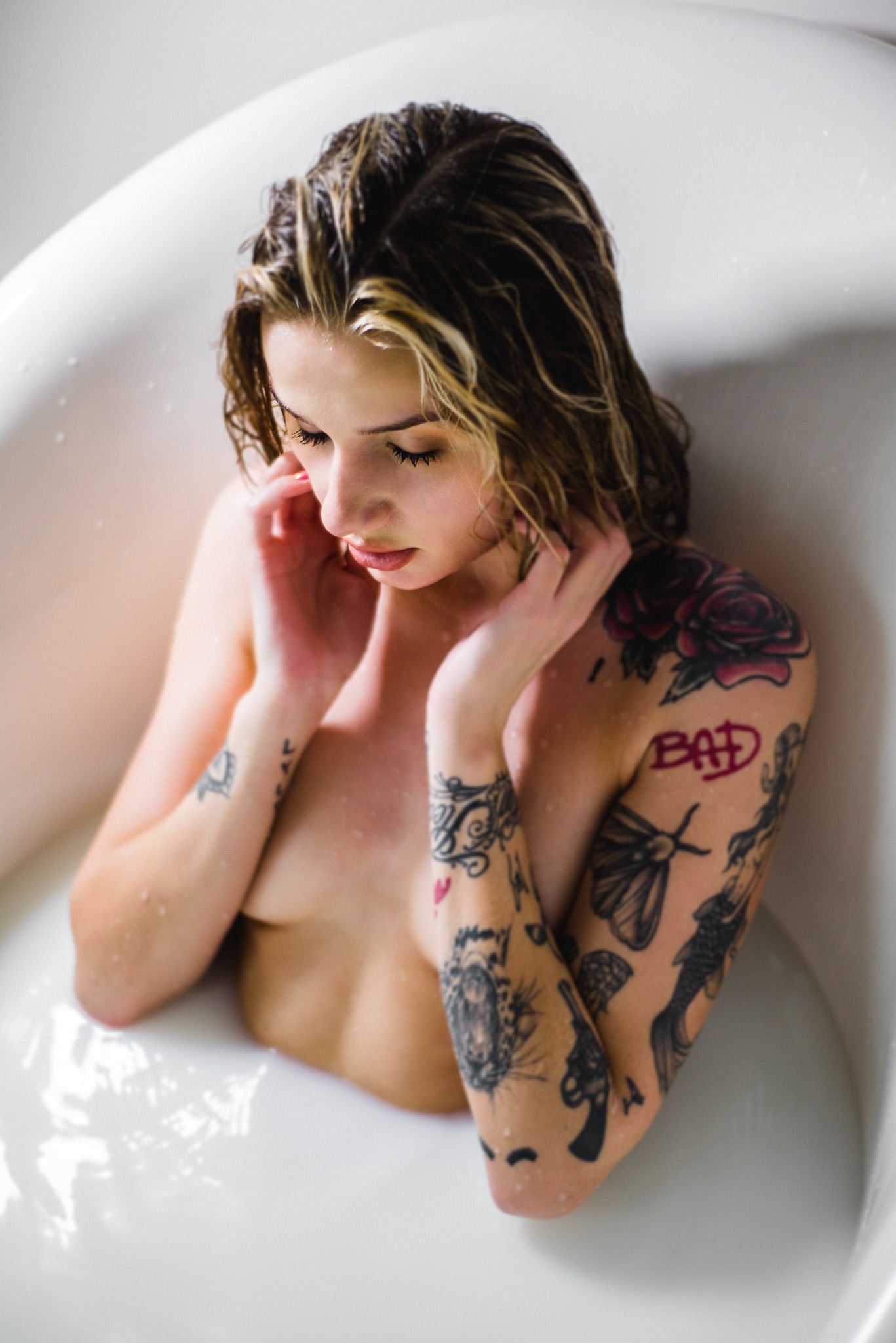 Kobieta z mokrymi włosami i tatuażami zanurzona w białej kąpieli, z delikatnym oświetleniem podkreślającym detale skóry i wzory na tatuażach.