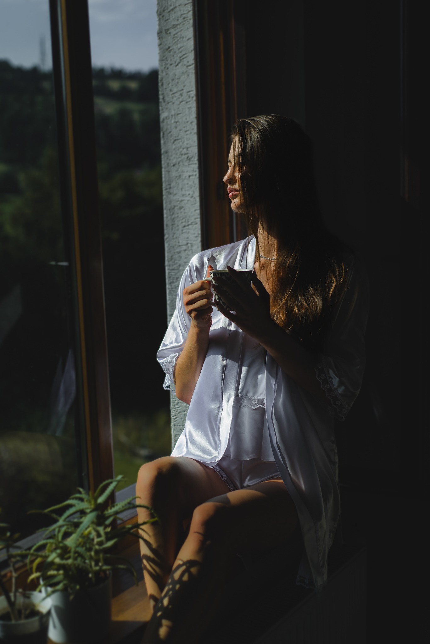 Kobieta w satynowej piżamie siedzi na parapecie, trzymając filiżankę i spoglądając przez okno na zalesione wzgórza w porannym słońcu.