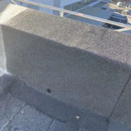 Fragment dachu pokrytego papą z widocznym kominem wentylacyjnym i elementami zabezpieczeń na dachu budynku przemysłowego w tle.