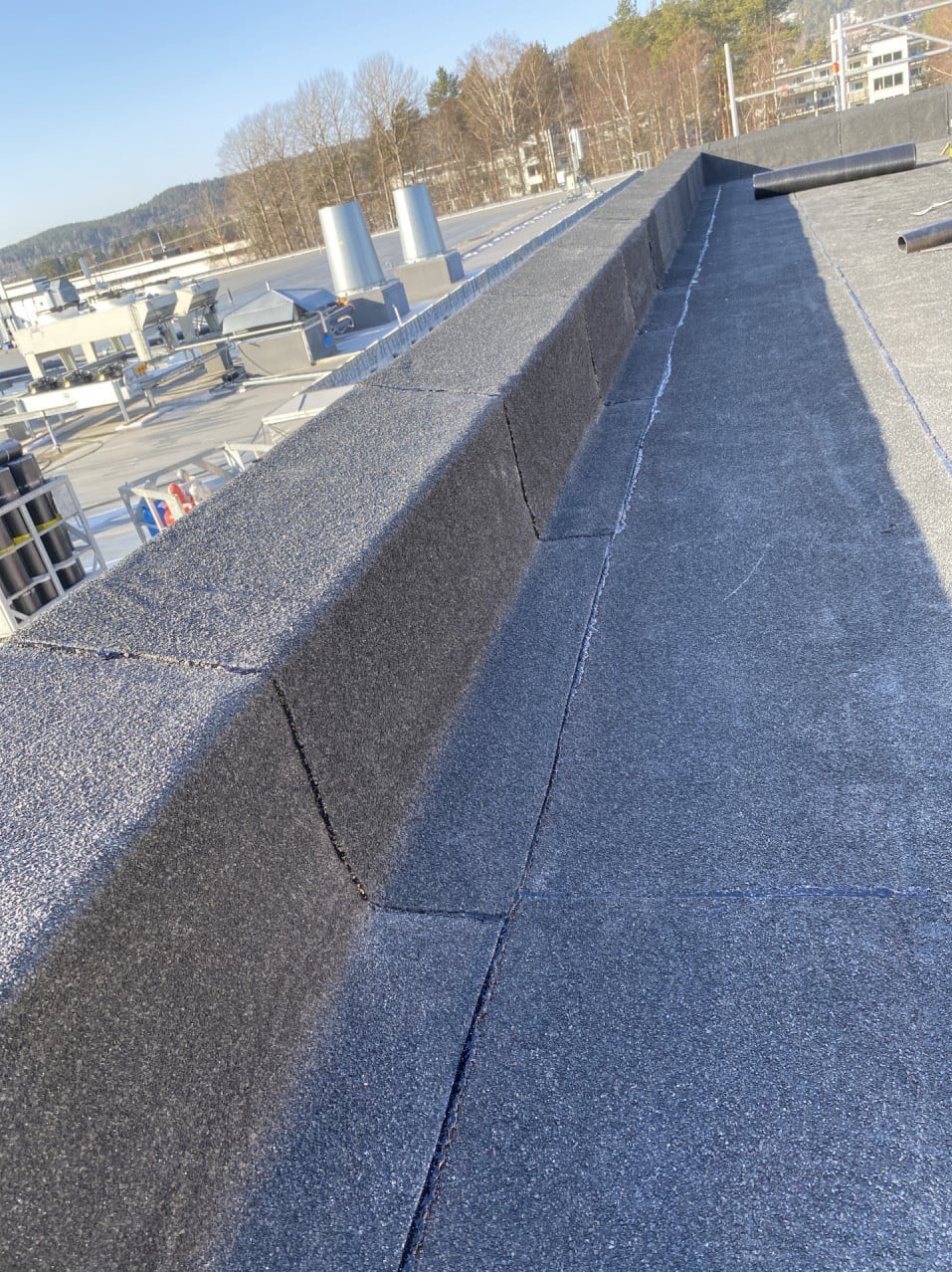 Detale izolacji dachu płaskiego z papy termozgrzewalnej, widoczne łączenia i obróbki blacharskie, w tle elementy instalacji przemysłowej i drzewa.