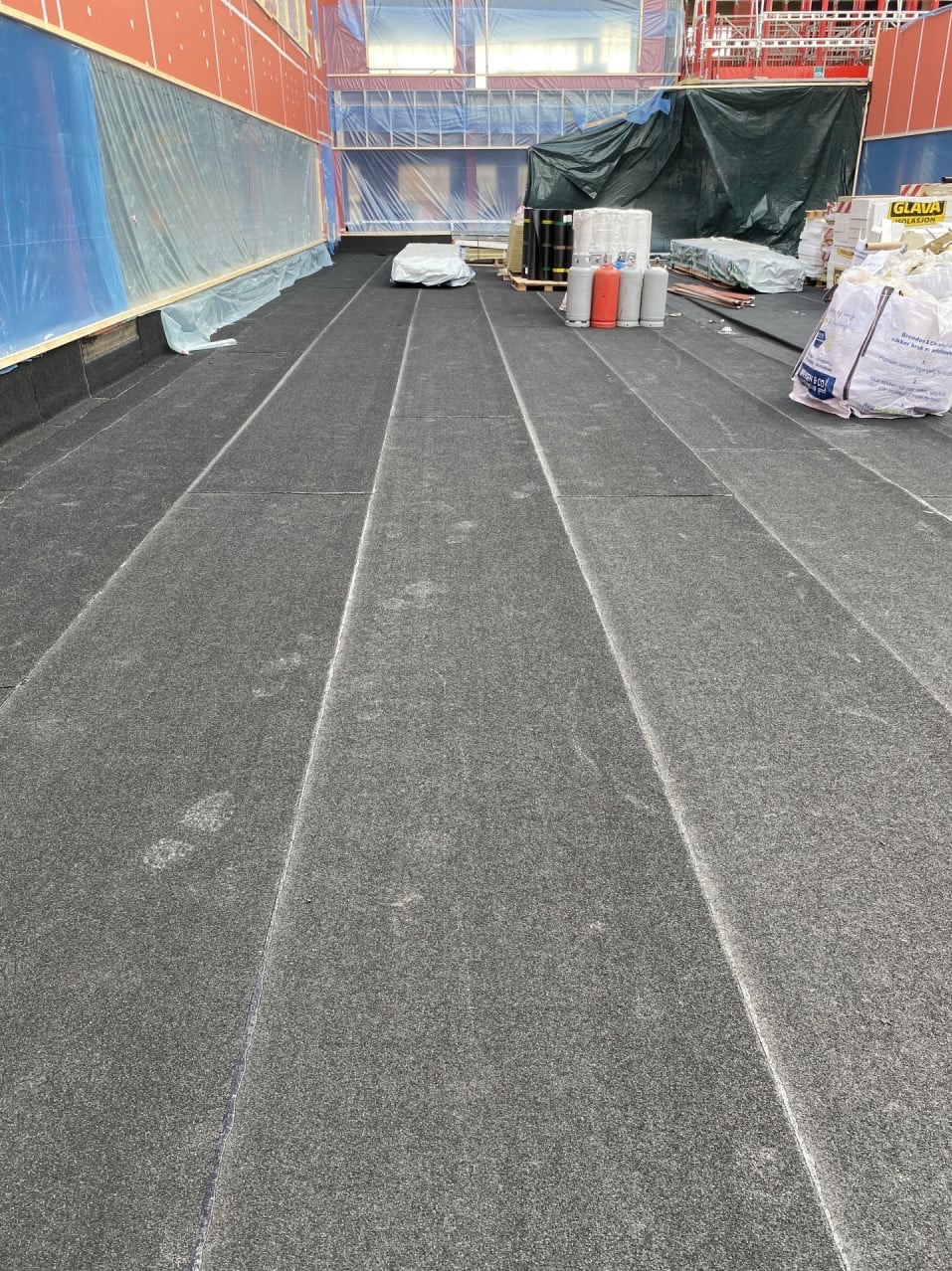 Powierzchnia dachu pokryta czarną papą w pasach, widoczne zgrzewy, w tle materiały budowlane, butle z gazem i zabezpieczenia rusztowania.