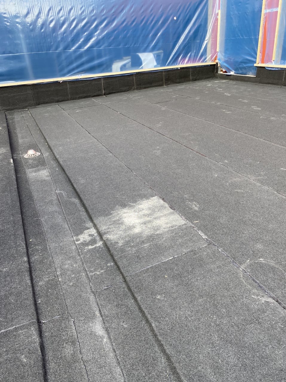 Widok z góry na płaski dach pokryty papą, z widocznym odpływem wody i zabezpieczeniem ścian folią budowlaną.