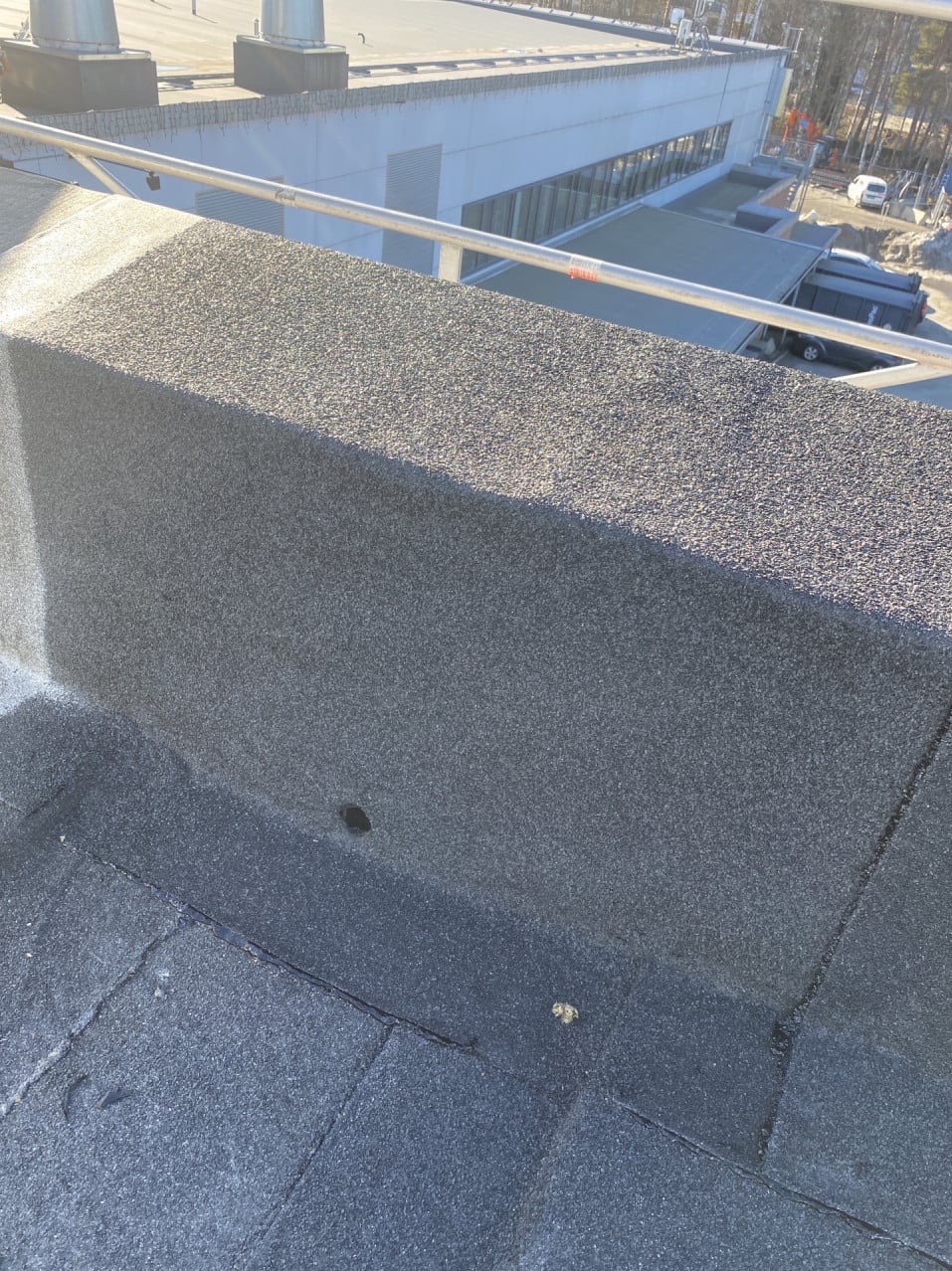 Fragment dachu pokrytego papą z widocznym kominem wentylacyjnym i elementami zabezpieczeń na dachu budynku przemysłowego w tle.