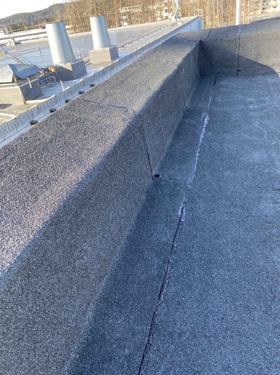 Widok z góry na płaski dach pokryty papą z widocznymi łączeniami i niewielkimi pęknięciami, w tle elementy instalacji technicznej budynku.