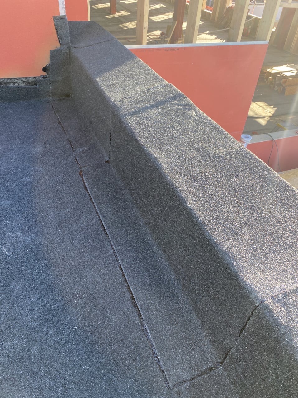 Szczegółowe ujęcie nowo położonej, czarnej papy na dachu z widocznymi łączeniami i fragmentami konstrukcji drewnianej w tle, oświetlone silnym światłem słonecznym.