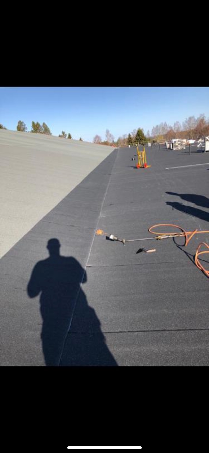 Prace dekarskie na płaskim dachu z użyciem palnika, widoczny cień osoby pracującej oraz narzędzia na tle krajobrazu.