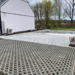 Powierzchnia częściowo wyłożona ażurowymi płytami betonowymi, dalej ułożona jasnoszara kostka, na drugim planie biały budynek z czarnym dachem i otoczenie z drzewami.