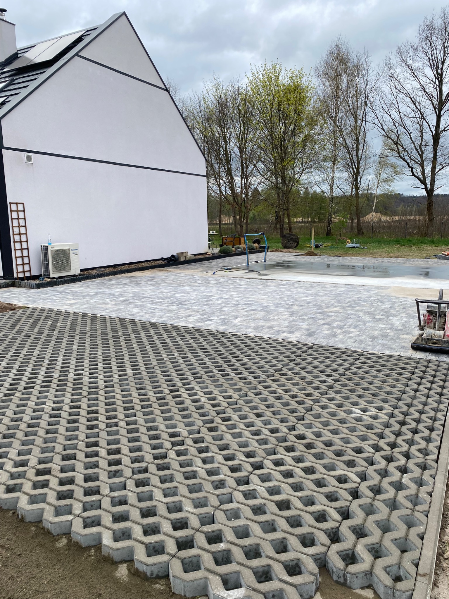 Powierzchnia częściowo wyłożona ażurowymi płytami betonowymi, dalej ułożona jasnoszara kostka, na drugim planie biały budynek z czarnym dachem i otoczenie z drzewami.