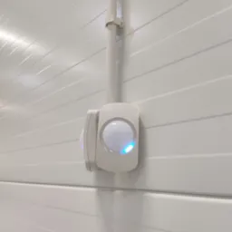 Biały czujnik ruchu zamontowany na białej, panelowej ścianie, z widoczną rurką instalacyjną i delikatnym, niebieskim światłem sygnalizacyjnym.