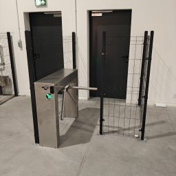 CAMLO Mateusz Górzyński - Metalowy, obrotowy kołowrót wejściowy z zieloną strzałką sygnalizującą przejście, otoczony czarnym ogrodzeniem z siatki, przed dwoma czarnymi drzwiami w hali przemysłowej.
