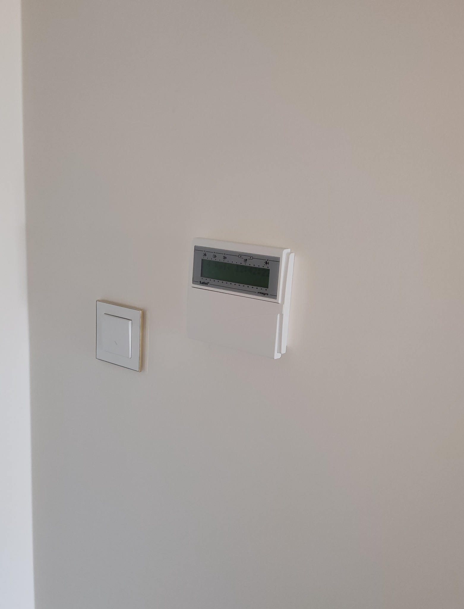 Zamontowany na ścianie panel sterowania alarmem marki Satel Integra, obok włącznik światła w ramce, na jednolitej, jasnej ścianie.