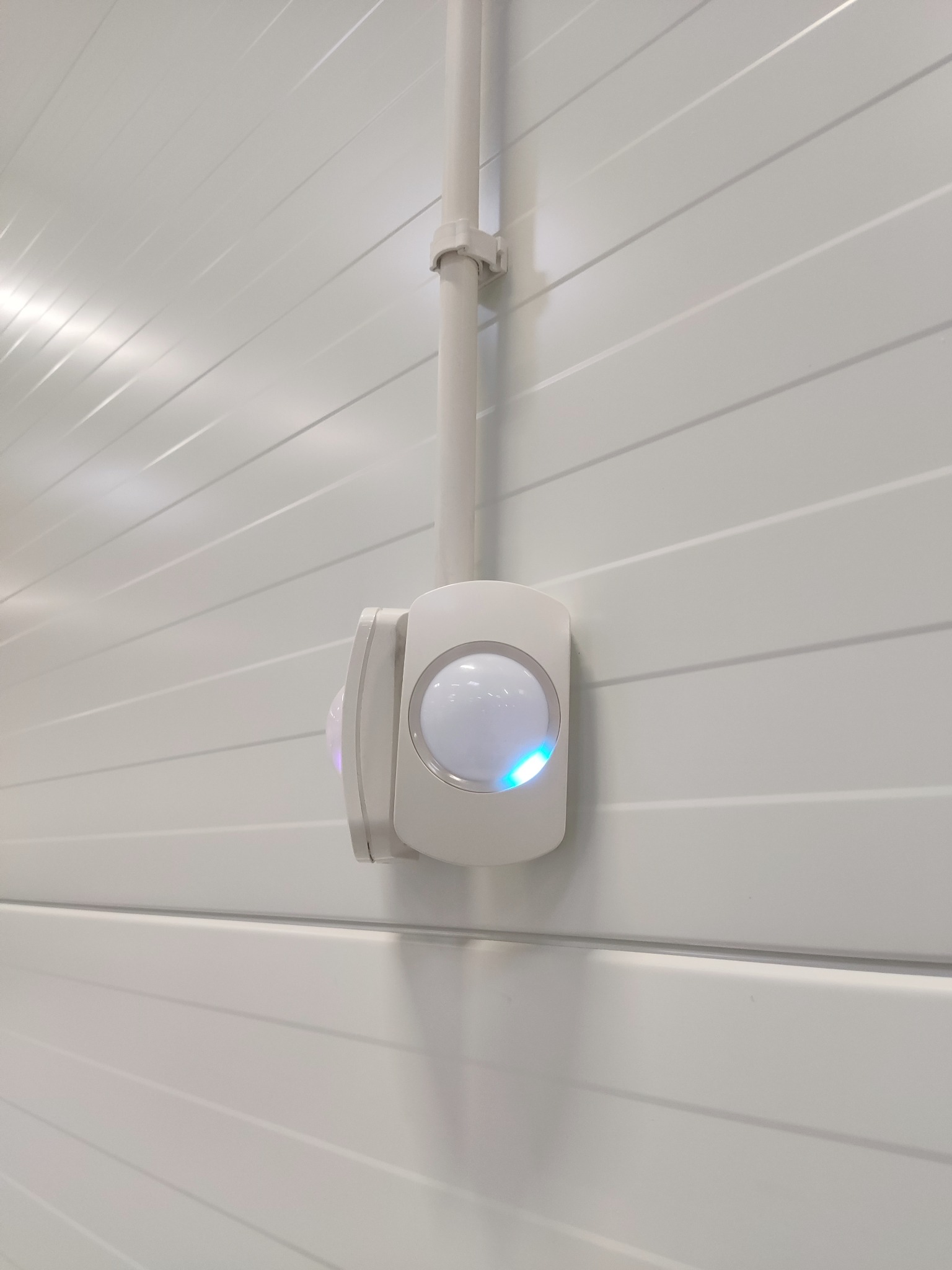 Biały czujnik ruchu zamontowany na białej, panelowej ścianie, z widoczną rurką instalacyjną i delikatnym, niebieskim światłem sygnalizacyjnym.