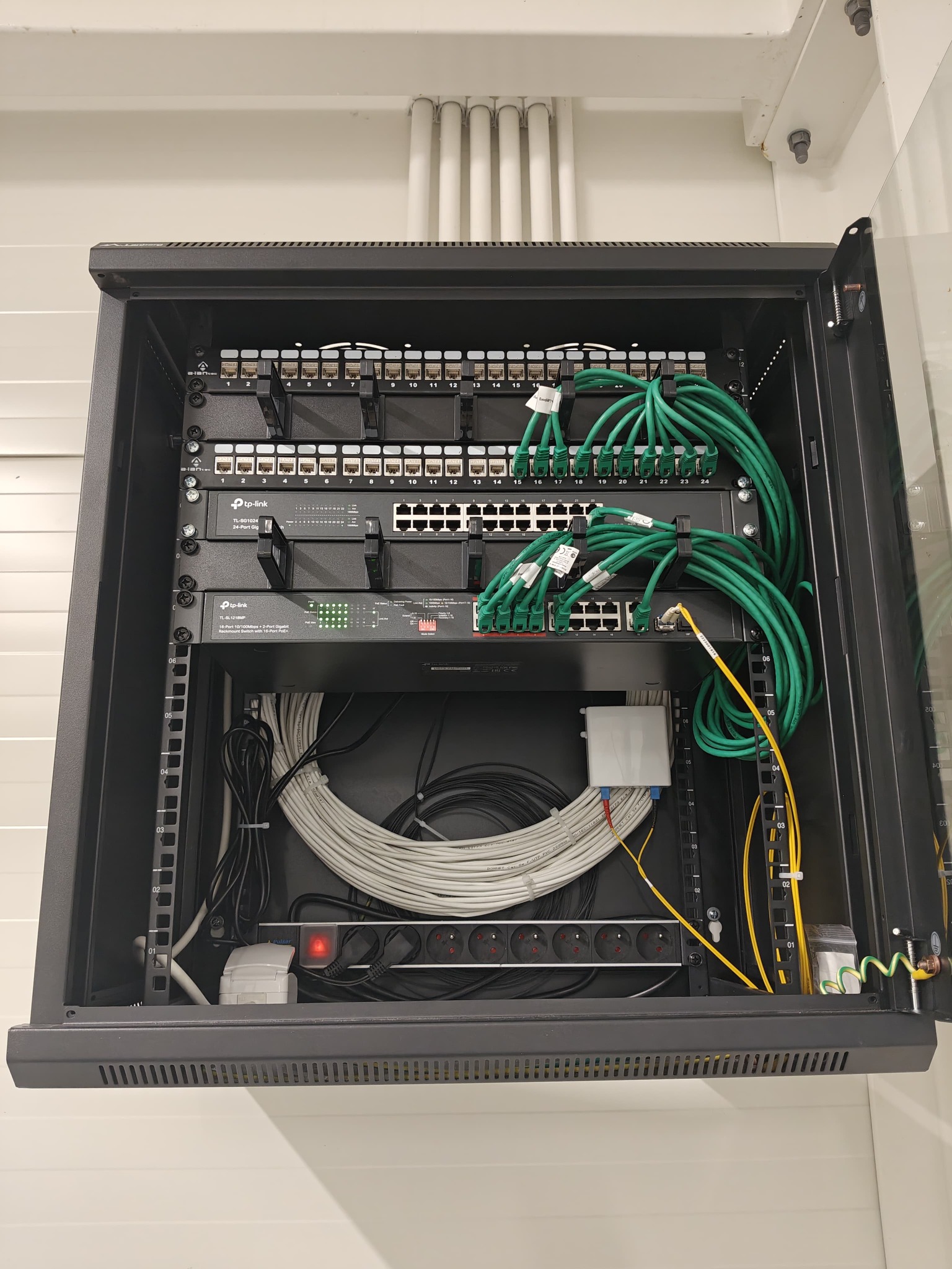 Szafa rack z przełącznikami sieciowymi TP-Link, patch panelem i uporządkowanymi kablami Ethernet w kolorach zielonym i białym, widoczne gniazda zasilające i uziemienie.