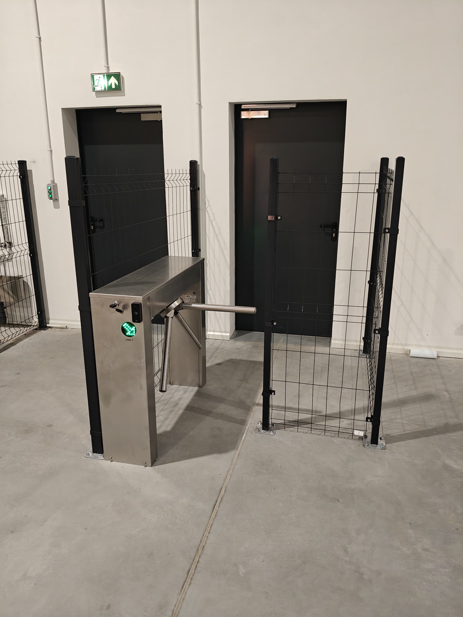Metalowy, obrotowy kołowrót wejściowy z zieloną strzałką sygnalizującą przejście, otoczony czarnym ogrodzeniem z siatki, przed dwoma czarnymi drzwiami w hali przemysłowej.
