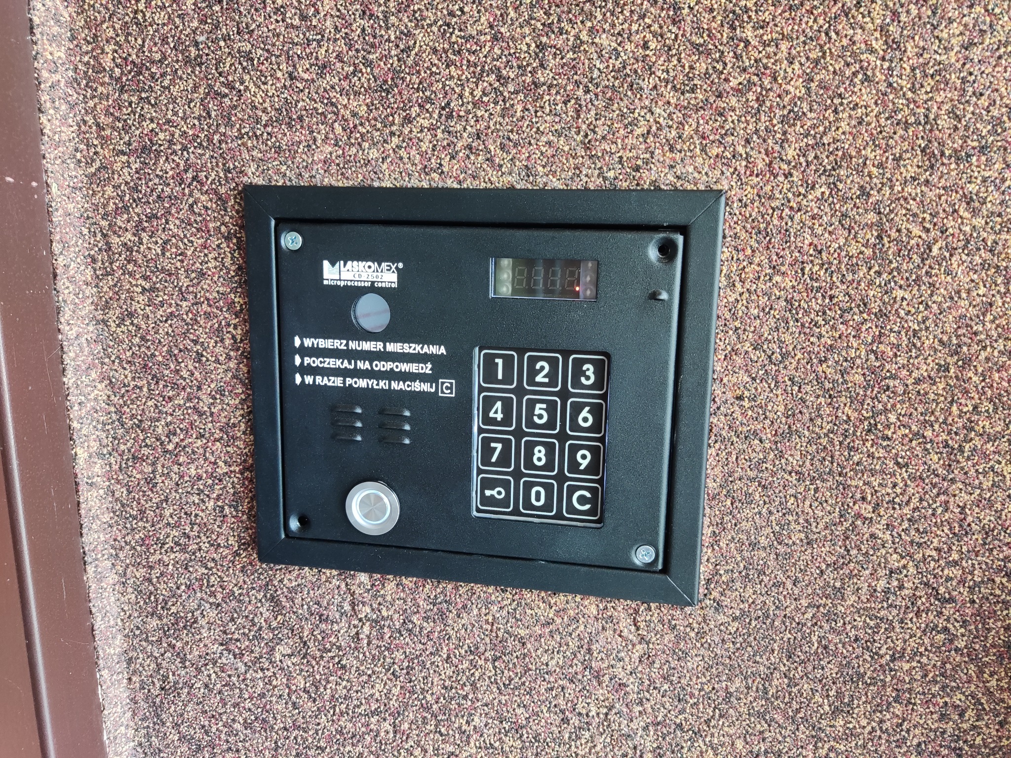 Panel domofonu Laskomex CD-2502 z klawiaturą numeryczną i wyświetlaczem LED, zamontowany na ścianie o chropowatej fakturze.