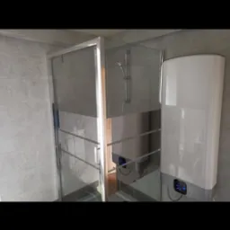 Wykończona łazienka z kabiną prysznicową ze srebrnymi ramami i dwoma białymi podgrzewaczami wody z niebieskimi wyświetlaczami na tle szarych płytek.