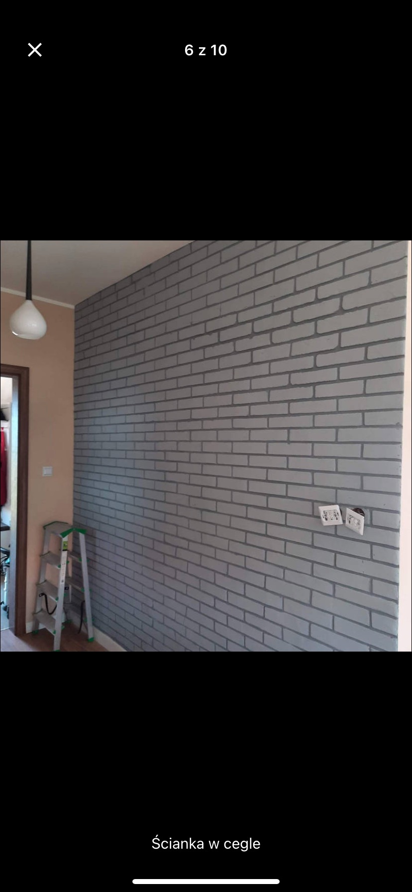 Ściana w pokoju wykończona farbą imitującą cegłę, widoczne gniazdka elektryczne i drabina malarska w rogu pomieszczenia.