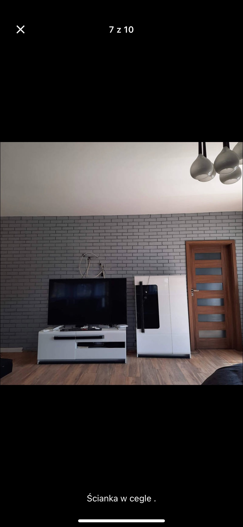 Szara ściana z cegły w salonie, z widocznymi kablami nad telewizorem, biała szafka RTV, drewniana podłoga i drzwi z przeszkleniami.