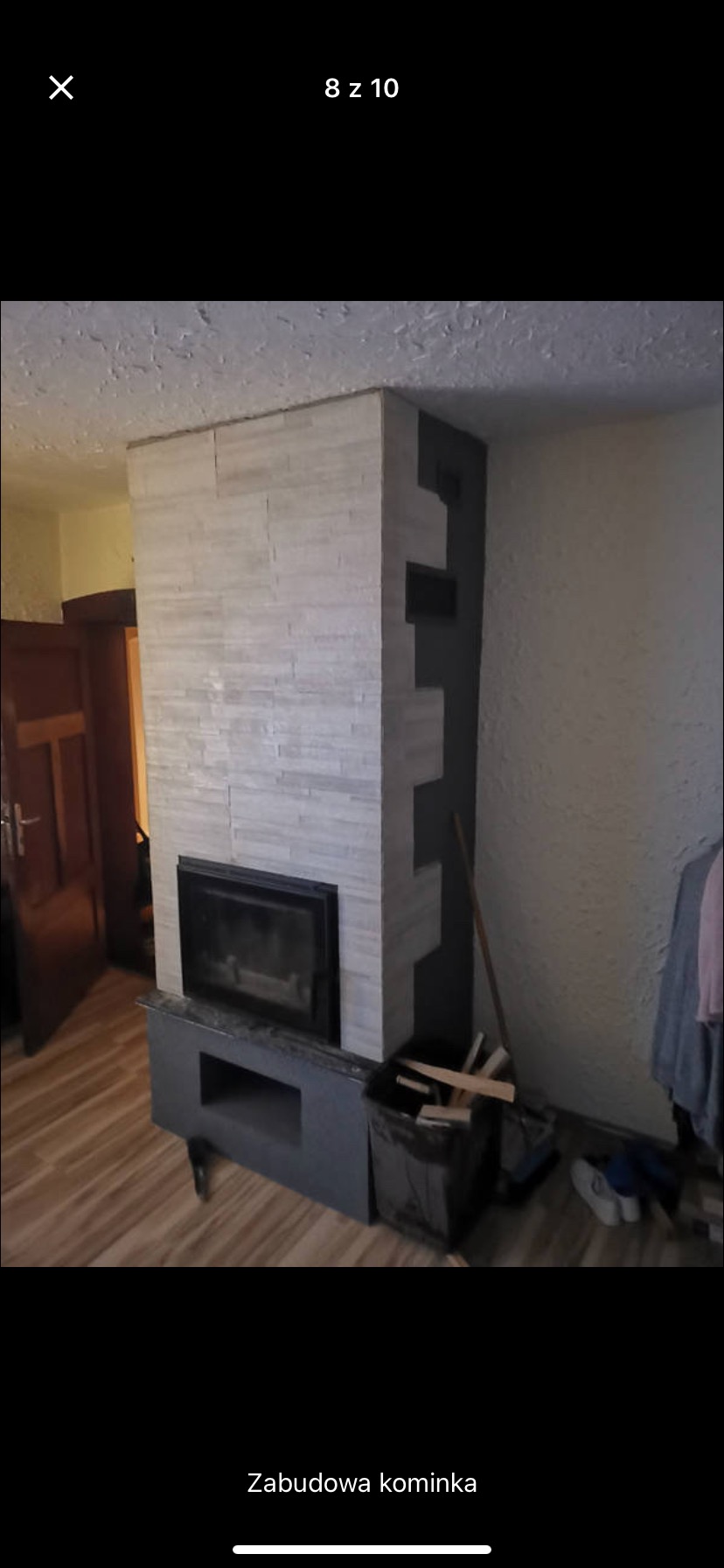 Obudowa kominka wykończona płytkami imitującymi kamień, z czarnymi elementami konstrukcyjnymi, widoczna w tle drewniana podłoga i drzwi.