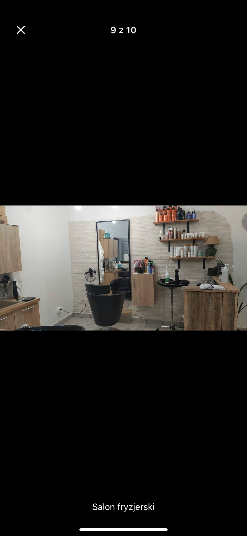 Wnętrze salonu z fotelem fryzjerskim, umywalką i drewnianymi półkami na kosmetyki, wykończone cegłą dekoracyjną.