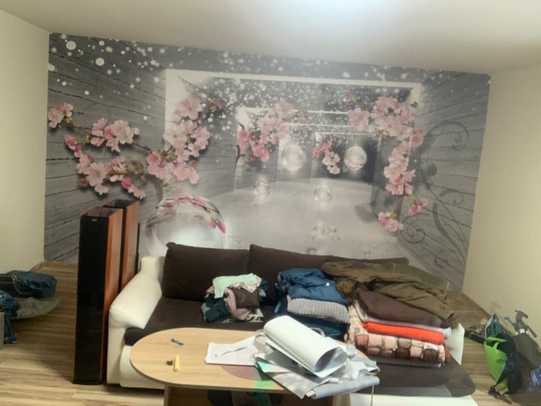 Pokój z fototapetą przedstawiającą kwitnącą wiśnię i perspektywę tunelu, z brązową sofą i stertą ubrań na stole.