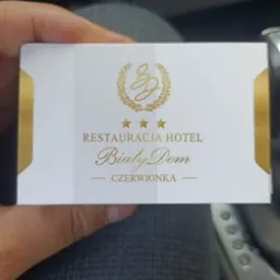 Biała wizytówka z logo złotego wieńca i napisem Restauracja Hotel Biały Dom Czerwionka, trzymana w dłoni.