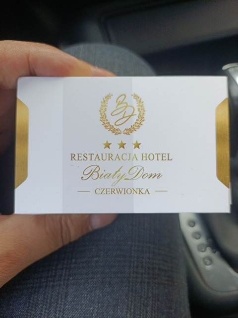 Biała wizytówka z logo złotego wieńca i napisem Restauracja Hotel Biały Dom Czerwionka, trzymana w dłoni.