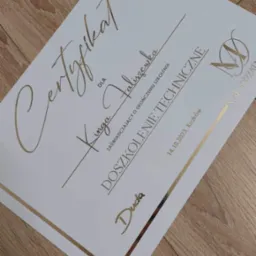 Elegancki certyfikat z elementami w kolorze złotym na białym tle, leżący na drewnianej powierzchni. Certyfikat potwierdza ukończenie doszkolenia technicznego przez Kingę Faliszewską.