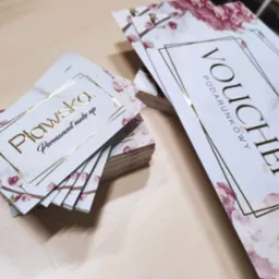 Stosy wizytówek i voucherów z motywem kwiatowym i złotymi napisami na jasnym drewnianym tle, skierowane ukośnie w prawą stronę.