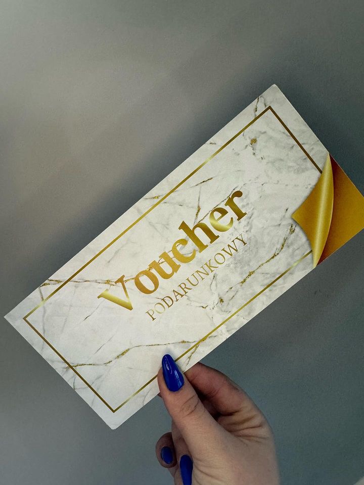 Dłoń z niebieskimi paznokciami trzyma elegancki voucher prezentowy z marmurkowym tłem i złotymi napisami 'Voucher Podarunkowy', z odklejonym złotym rogiem.