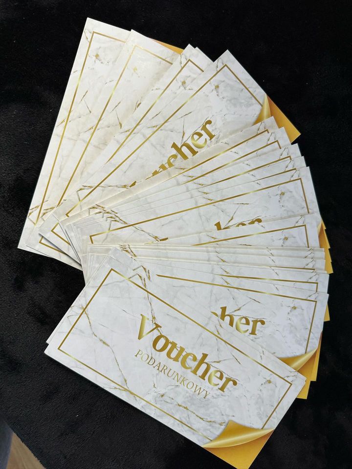 Stos eleganckich, marmurkowych voucherów prezentowych z złotymi ramkami i napisem 'Voucher Podarunkowy' na czarnym tle.