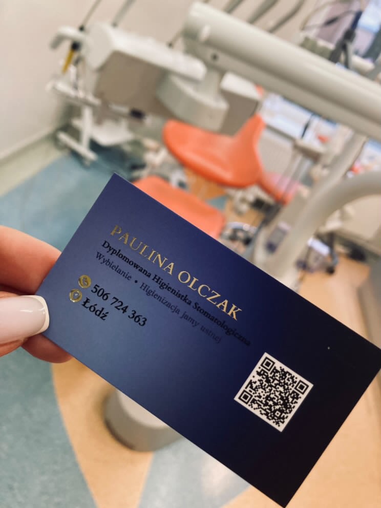 Granatowa wizytówka z złotym napisem 'Paulina Olczak Dyplomowana Higienistka Stomatologiczna' trzymana w dłoni na tle rozmytego gabinetu stomatologicznego z fotelem dentystycznym.
