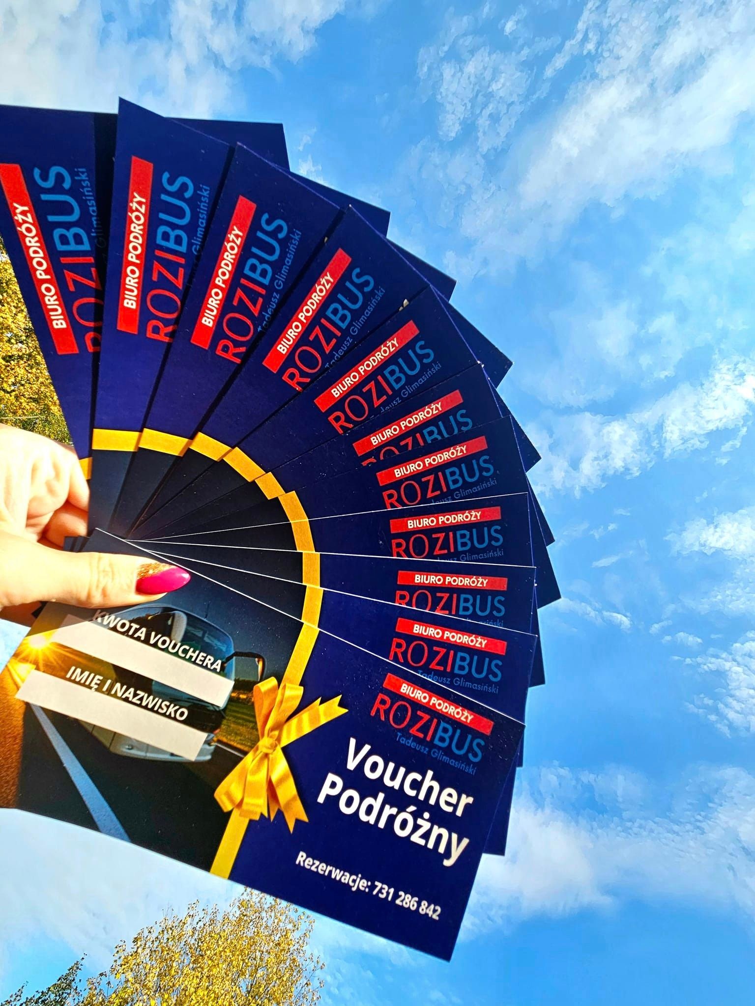 Dłoń trzymająca wachlarz błękitnych voucherów podróżnych z logo biura podróży, na tle błękitnego nieba i drzew
