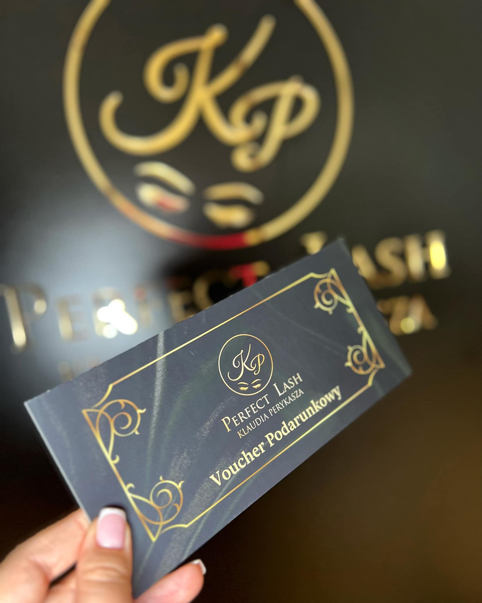 Dłoń trzymająca elegancki, granatowy voucher podarunkowy z złoconymi zdobieniami i logo 'Perfect Lash Klaudia Perykasza', widoczne w tle złote logo firmy na czarnym tle.